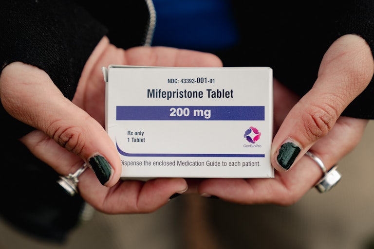 Hands with silver rings and black nail polish hold a small box that reads "Mifepritone Tablet 200 mg"